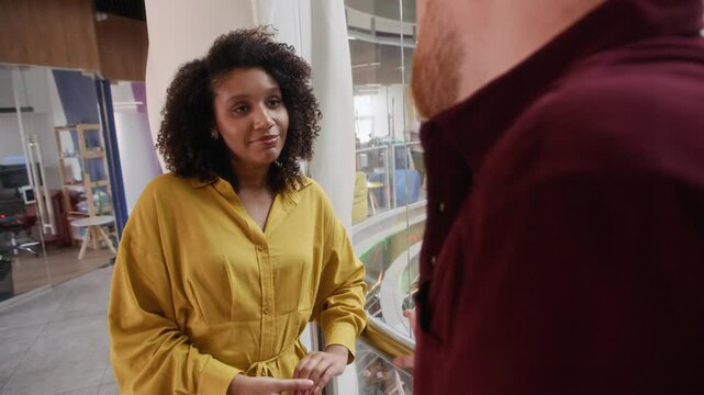 Medium handheld shot of bicultural gen Z coworkers chatting in hall of modern office building