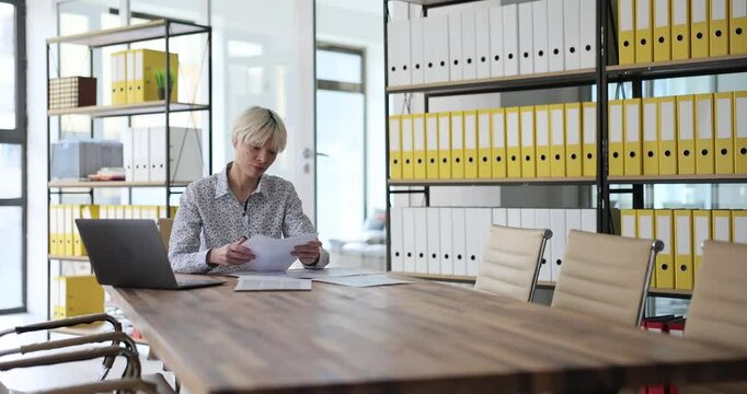 Woman with short hair sitting at long wooden conference table reviews documents. Focused preparation for board meeting in organized office environment