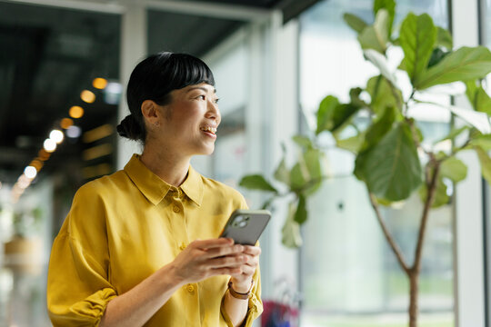 Smiling woman in yellow shirt texting on phone in modern office