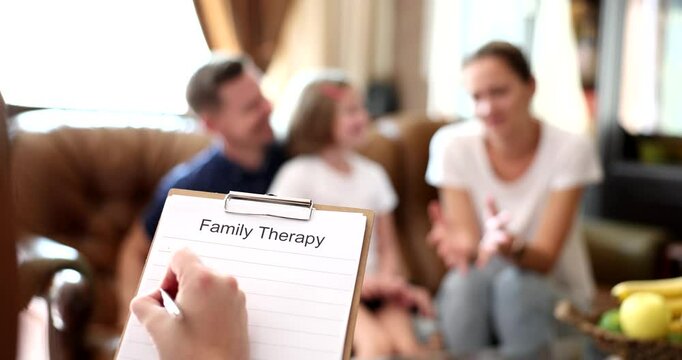 Family sits together on couch talking with therapist holding clipboard. Group engages in open dialogue building emotional understanding slow motion