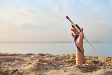 Young female hand holding paintbrush emerges from sandy beach. Serene seascape, highlighted by soft...