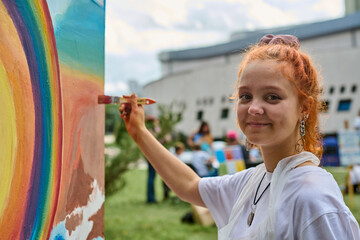 Young female with red hair paints vivid mural outdoors, smiling warmly. Green park, clear sky, vibrant colors create lively atmosphere. Casual attire, relaxed mood