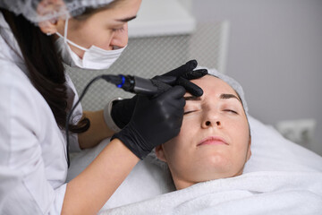 Beautician applies permanent makeup to young woman eyebrows in salon. Focus is on precision and...