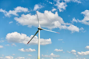 Modern wind turbine with red tipped blades stands tall against vivid blue sky filled with scattered white clouds, reflecting blend of technology and natural harmony