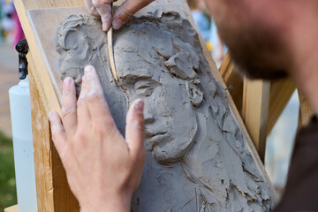 Adult man uses wooden tools, sculpting clay relief portrait. Natural light highlights textures....