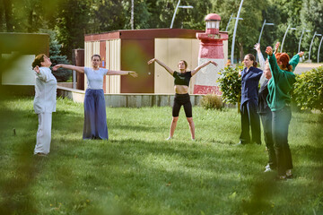 Group of adults practicing qigong in grassy garden. Female teacher leading session under sunny sky....