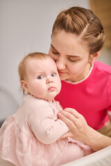 Young mother with brown hair holds baby girl in pink dress indoors. Warm lighting creates intimate, cozy environment. Baby curious expression contrasts with mothers gentle smile
