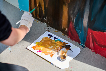 Focused view of adults hand painting mural outdoors. Gloved hand holds brush applying vivid red and...