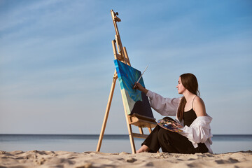 Young female artist paints on beach, capturing serene ocean view. Setting evokes tranquility and...