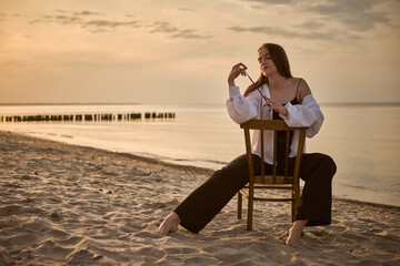 Young woman, styled as painter, sits contemplatively on chair at beach during sunset. Serene...