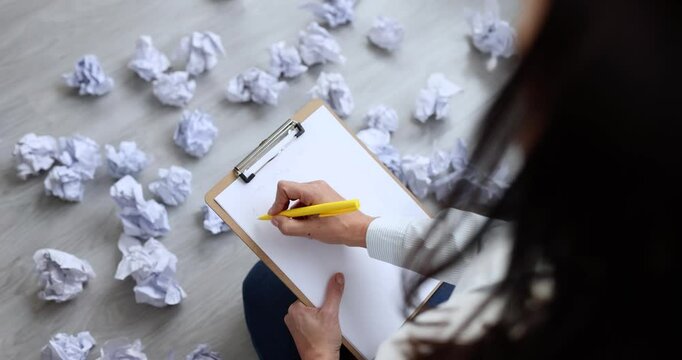 Woman leans over clipboard writing new draft among crumpled papers thrown on floor. Pencil presses sheet creating tension of mental rewriting struggle