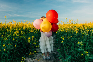 Girl in a white dress holding colorful balloons in a rapeseed field