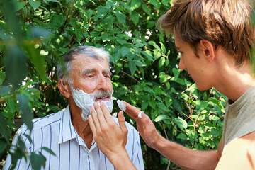 A young man helps an elderly man apply shaving foam on his face outdoors.