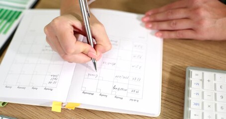 Woman holds pen and actively writes in planner displaying grid layout for monthly planning. Documents suggest organization and budgeting activities