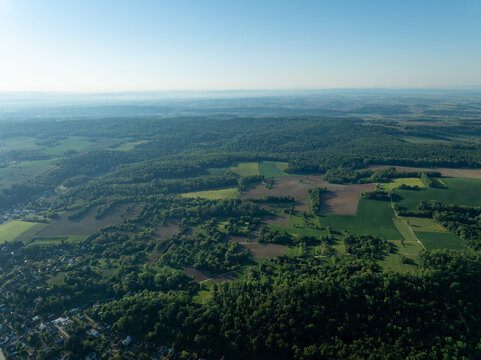 Aerial view of a dense, vibrant forest interspersed with agricultural fields, creating a tapestry of green and brown under a clear sky, Zillisheim, Grand Est, France.