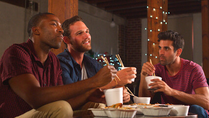 Group Of Male Friends Sharing Chinese Takeaway Meal And Watching TV In Loft Apartment