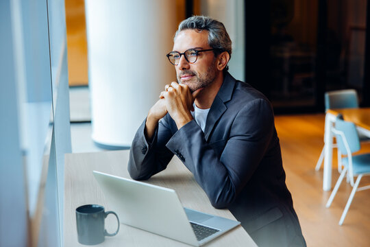 Thoughtful businessman sitting at desk by window with laptop and mug - Powered by Adobe