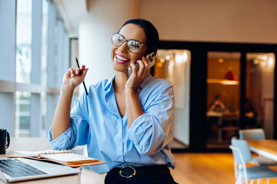 Smiling businesswoman talking on phone at desk with laptop and notes - Powered by Adobe