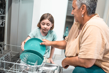 Child helps grandmother to load dishwasher. Family everyday life concept.