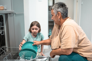 Child helps grandmother to load dishwasher. Family everyday life concept.