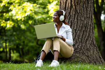 Happy black African woman sitting in the park and enjoying the nature. Listening to music and...