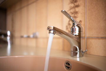 Shiny, chrome tap with running water stream inside apartment bathroom.