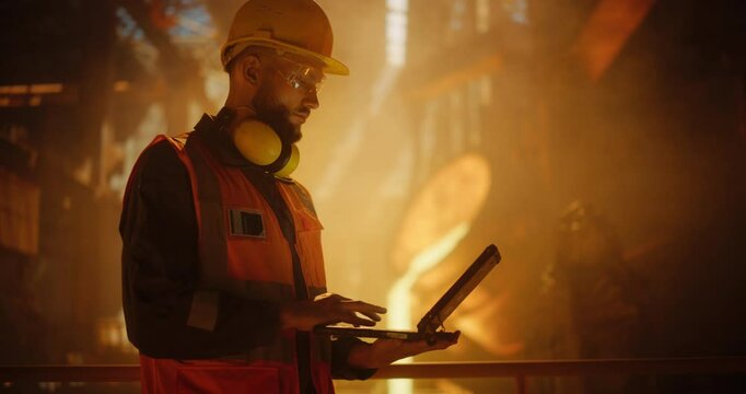 Caucasian Engineer Wearing High-Visibility Vest and Hard Hat, Working on a Laptop Computer in a Modern Foundry. Man Looking at Camera and Smiling. Furnace Crew Handles Molten Iron in the Background