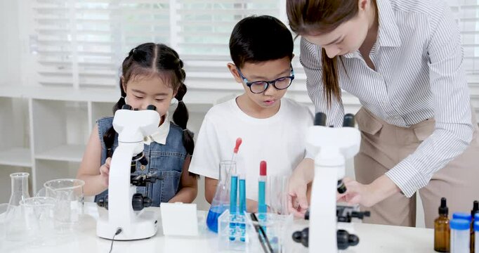 Asian female teacher teaching elementary students to operate microscope in science classroom lab while young boy adjusts settings and girl watches with interest and curiosity on her face