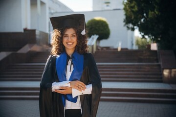 Feeling good, holding diploma. Cheerful graduate student in a gown is standing outdoors