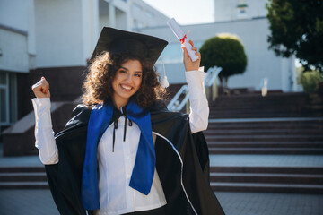 Diploma in hand. Cheerful graduate student in a gown is standing outdoors