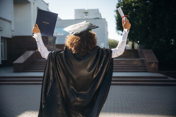Joyful, with diploma. Cheerful graduate student in a gown is standing outdoors