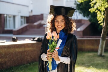 Gift of flowers in hands. Cheerful graduate student in a gown is standing outdoors