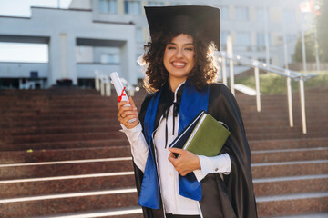 University building behind. Cheerful graduate student in a gown is standing outdoors