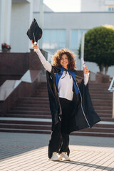 Cheerful graduate student in a gown is standing outdoors