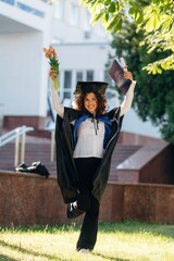 Green tree and university building behind. Cheerful graduate student in a gown is standing outdoors