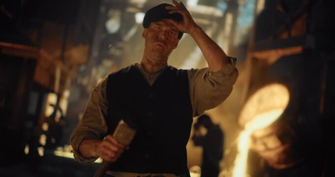 Working-Class Man of the Industrial Era Picking Up a Sledgehammer in Front of Camera, Taking His Cap Off and Wipes Away Sweat From His Forehead. Labor Worker Standing in Front of an Iron Smelter