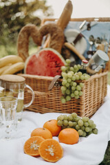A rustic picnic setup with wicker basket, fresh fruits like grapes, oranges, watermelon, baguette, and glassware on a white tablecloth. Cozy summer or countryside picnic concept.