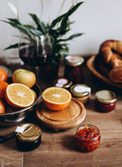 Cozy rustic breakfast table with fresh bread, citrus fruits, and assorted jam jars, half of orange and red wine glass 