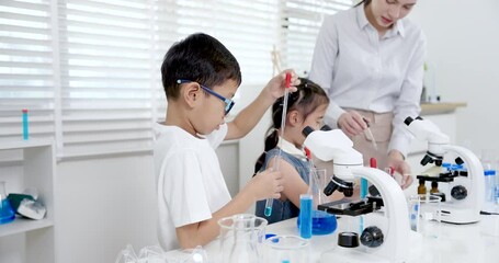 Asian boy using pipette to drop blue solution into testtube while young Asian girl observing experiment beside microscope during interactive primary science class supervised by Asian female teacher - Powered by Adobe