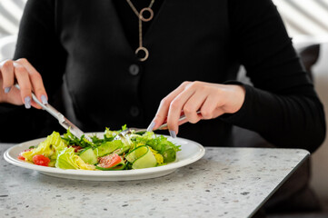 Woman in black outfit eating fresh vegetable salad with fork and knife, close-up of hands at modern kitchen table, promoting healthy lifestyle and balanced nutrition.