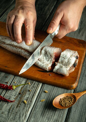Hands are slicing a fresh fish on a wooden surface. Nearby, dried chilies and a spoon of coriander seeds create a rustic cooking atmosphere in a warm kitchen