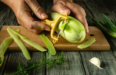 A pair of hands expertly peels a striped vegetable marrow or squash using a yellow peeler while surrounded by fresh herbs and a garlic clove on a wooden surface