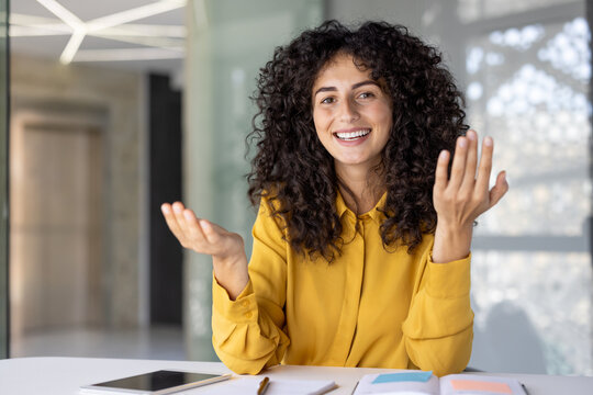 A cheerful woman with curly hair, gestures expressively while smiling, possibly presenting or explaining something in an office setting.