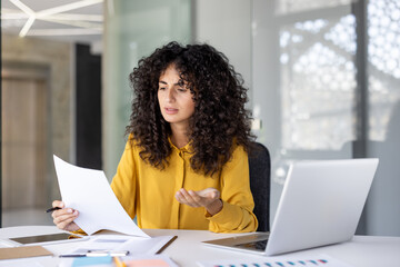 A puzzled woman in a yellow blouse is reviewing documents in an office setting, with a laptop...
