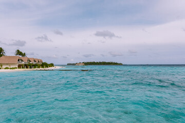 Scenic view of a tropical coastline with clear turquoise waters and distant island villas under a cloudy sky. A perfect representation of peaceful seascapes and luxury island life in paradise.
