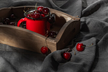 A wooden tray and a red mug with ripe cherries on a gray tablecloth.