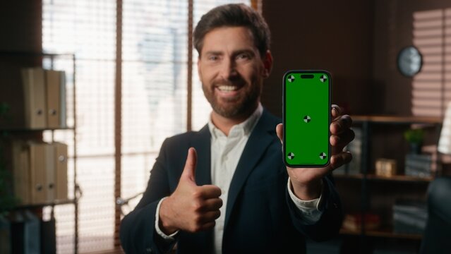 Smiling happy middle aged business man showing green screen mobile phone like gesture thumb up holding smartphone chroma key display success achieve mature businessman male looking at camera in office