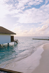 Gentle ocean waves caress the empty tropical shore at sunrise, with an overwater bungalow casting...