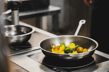 Sautéed vegetables including broccoli, potatoes, and yellow bell peppers cooking in a stainless...