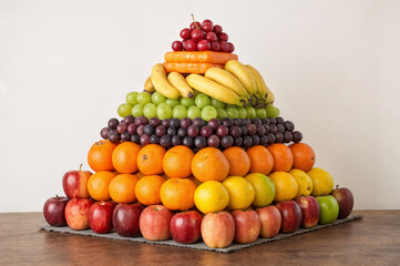 shows a pyramid-shaped arrangement of various fruits, including bananas, grapes, oranges, apples, and lemons, placed on a wooden table. The fruits are stacked in a colorful, rainbow-like manner, with 